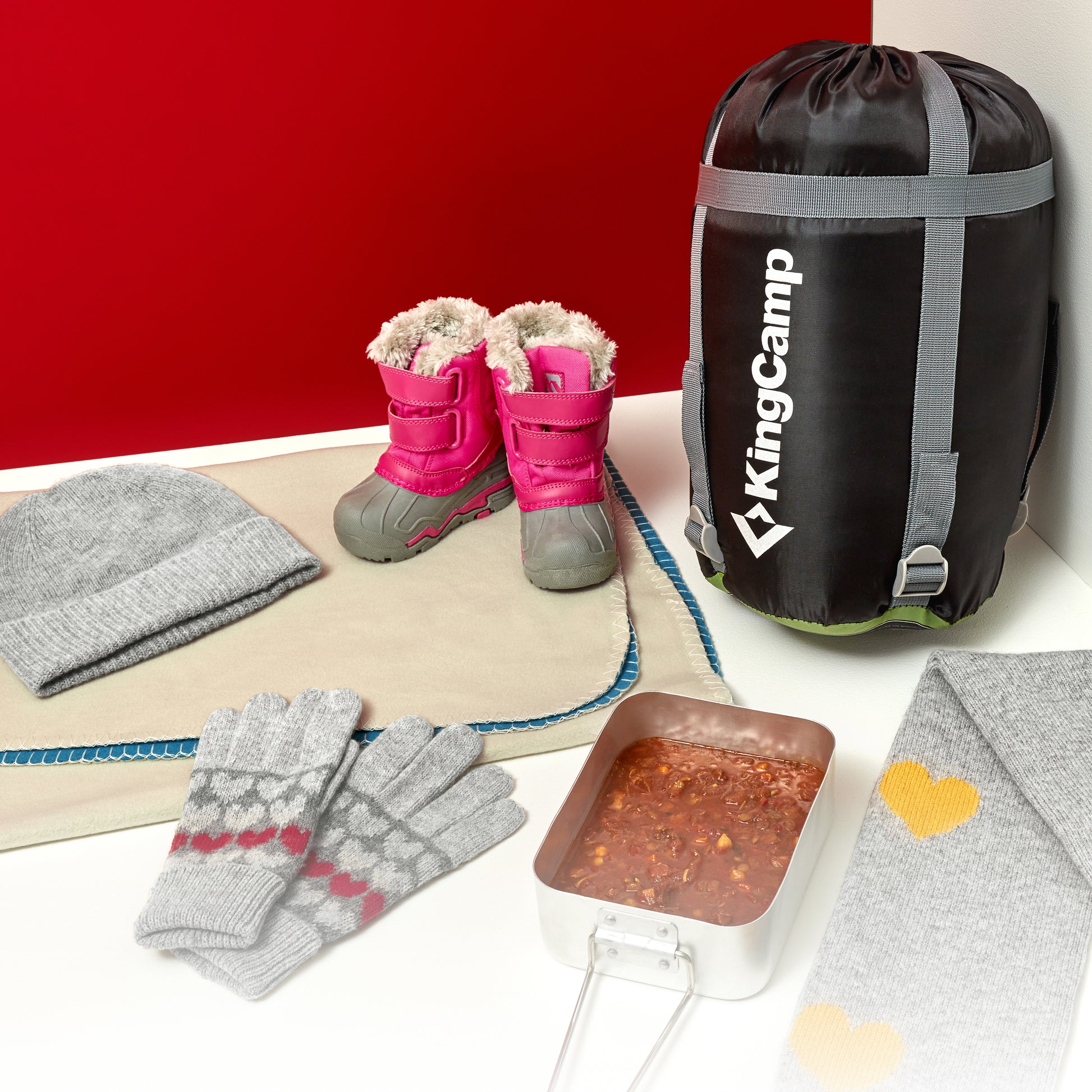 In the image there is a sleeping bag packed in a sack from KingCamp, a pair of girls winter boots, a blanket, a pair of winter essentials such as gloves, hat and scarf, and a portion of hot food in a metal container.