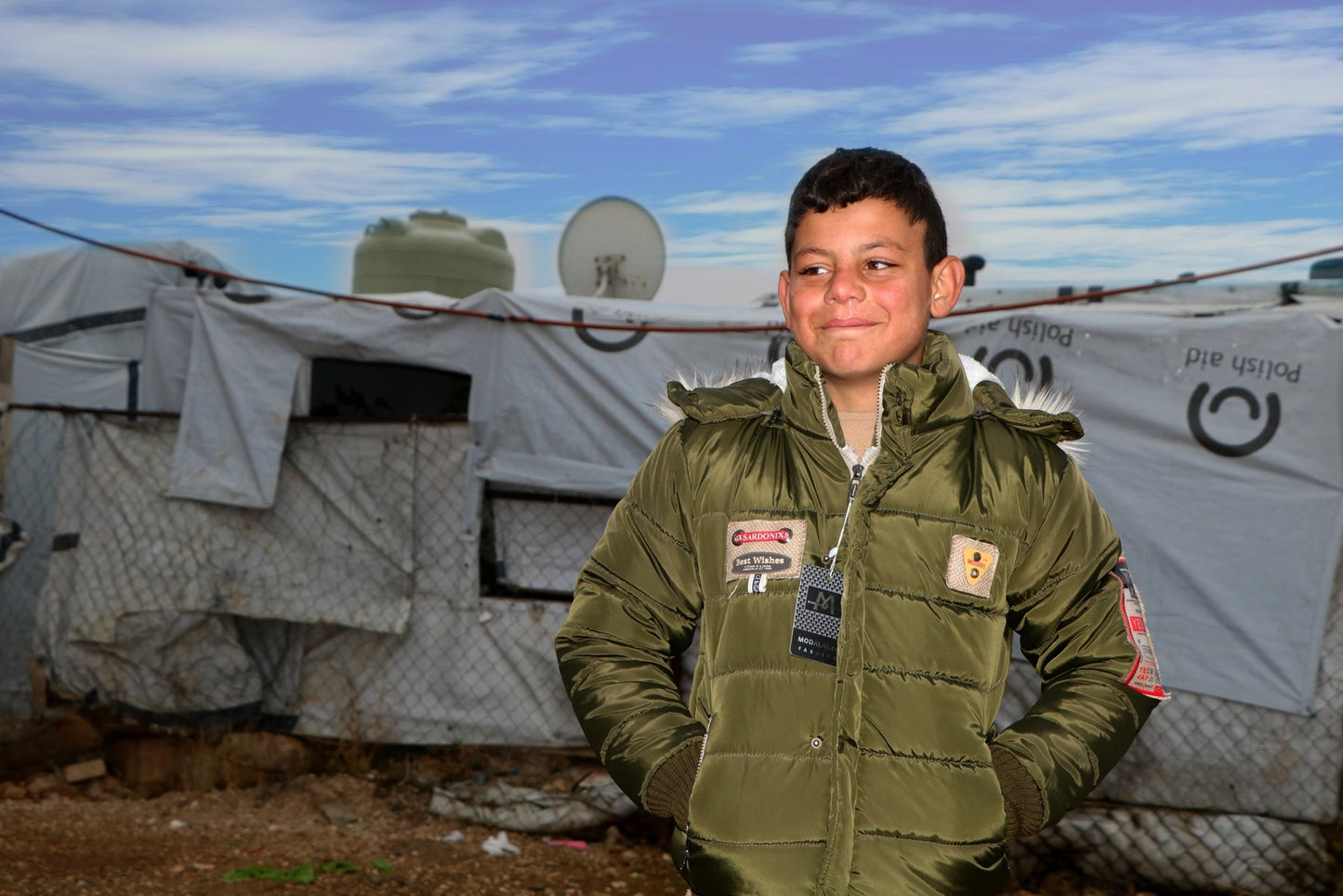 A young boy dressed for winter, in a refugee camp. Behind there is improvised accommodation covered by plastic sheets.