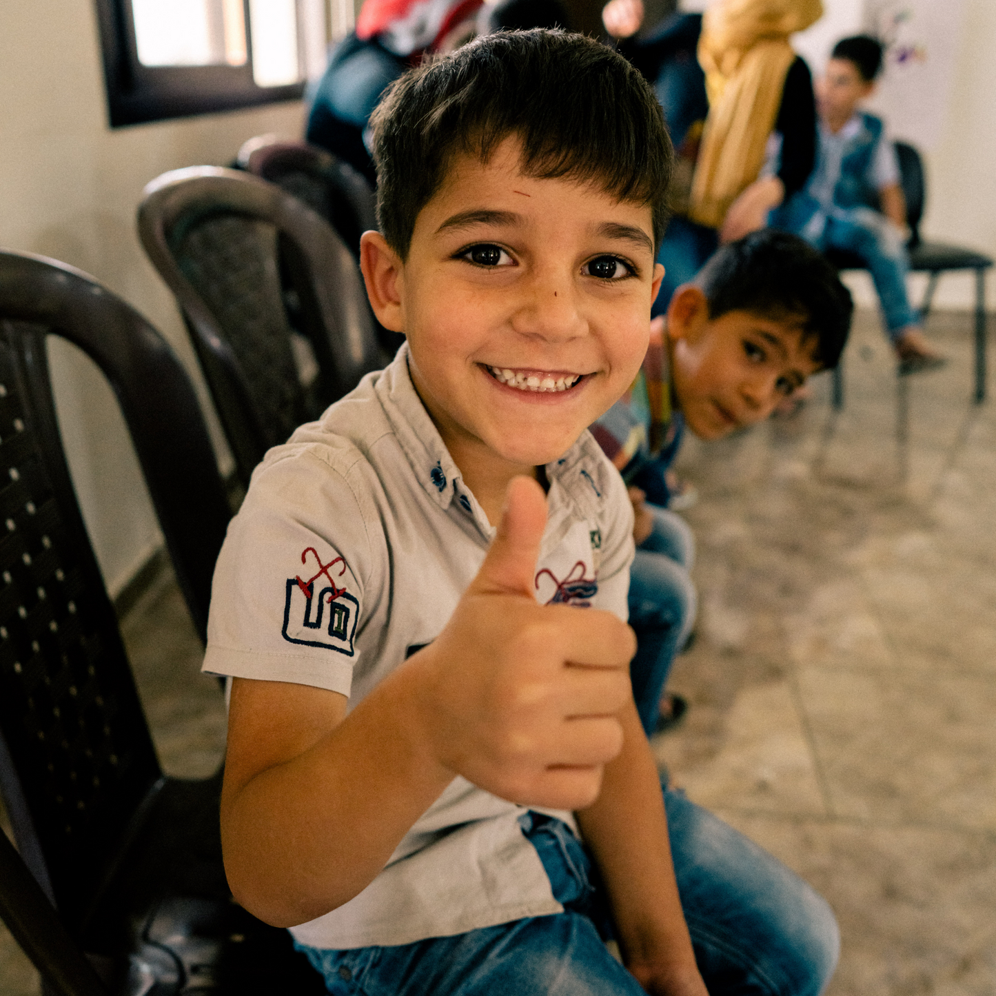 This is an image taken in a room with children refugees who are sitting down on chairs and socialising, and the image has a focus on one child who is smiling and showing thumbs up.